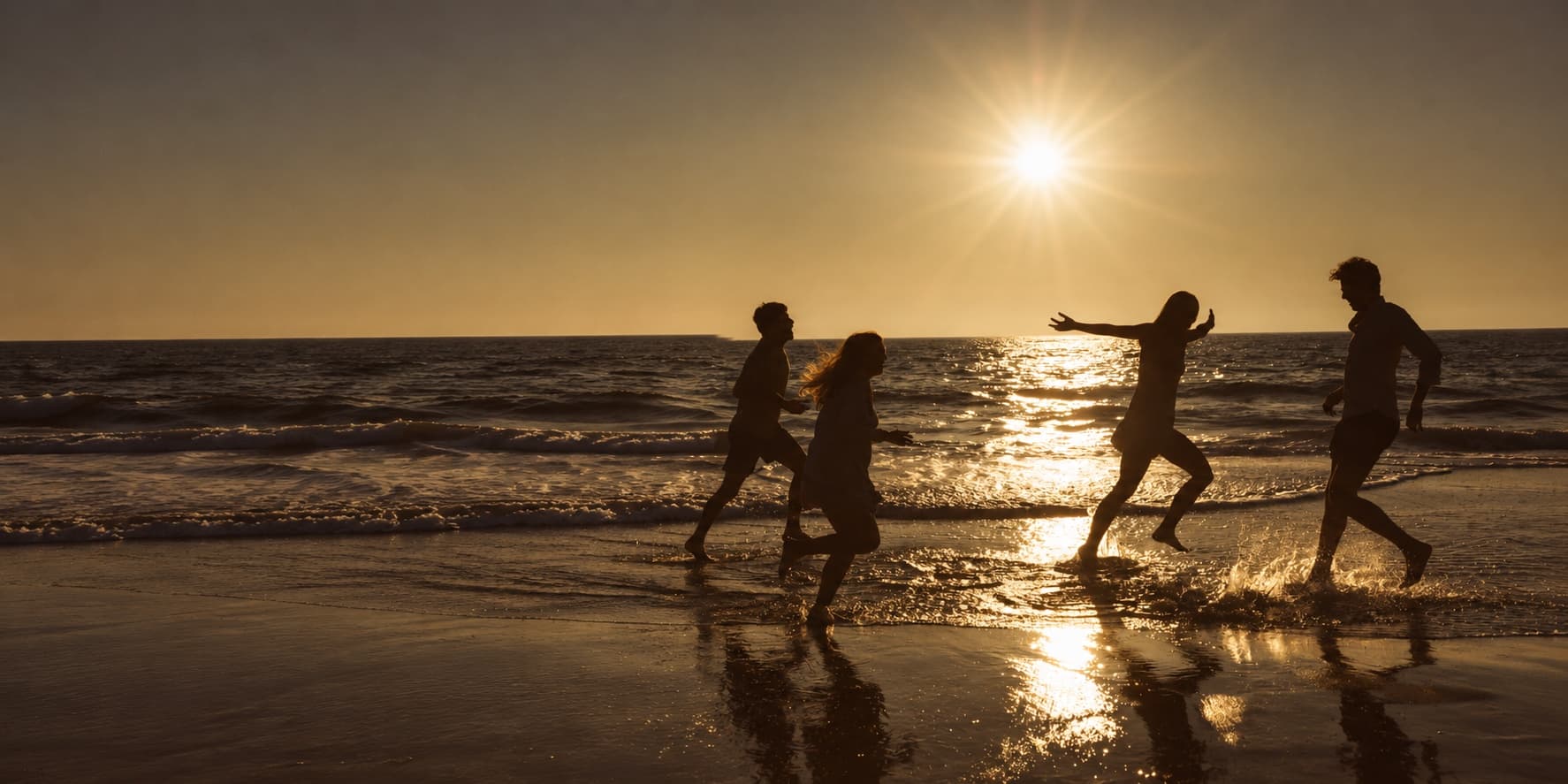 Four friends playing together in the ocean at sunset, with arms outstretched and water splashing around them